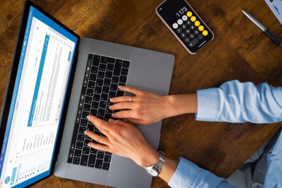 A person types on a laptop keyboard with a calculator and pen nearby, focusing on a digital task on the screen.