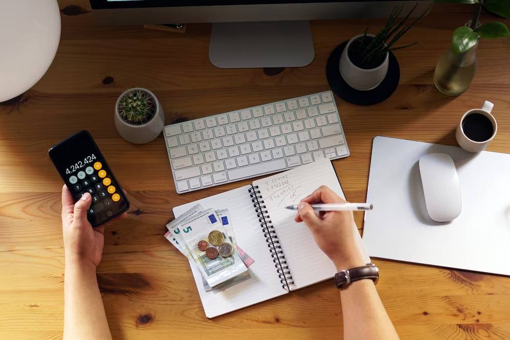 A person in a gray sweater takes notes from an open book while using a laptop, surrounded by a phone, calculator, and notebooks.
