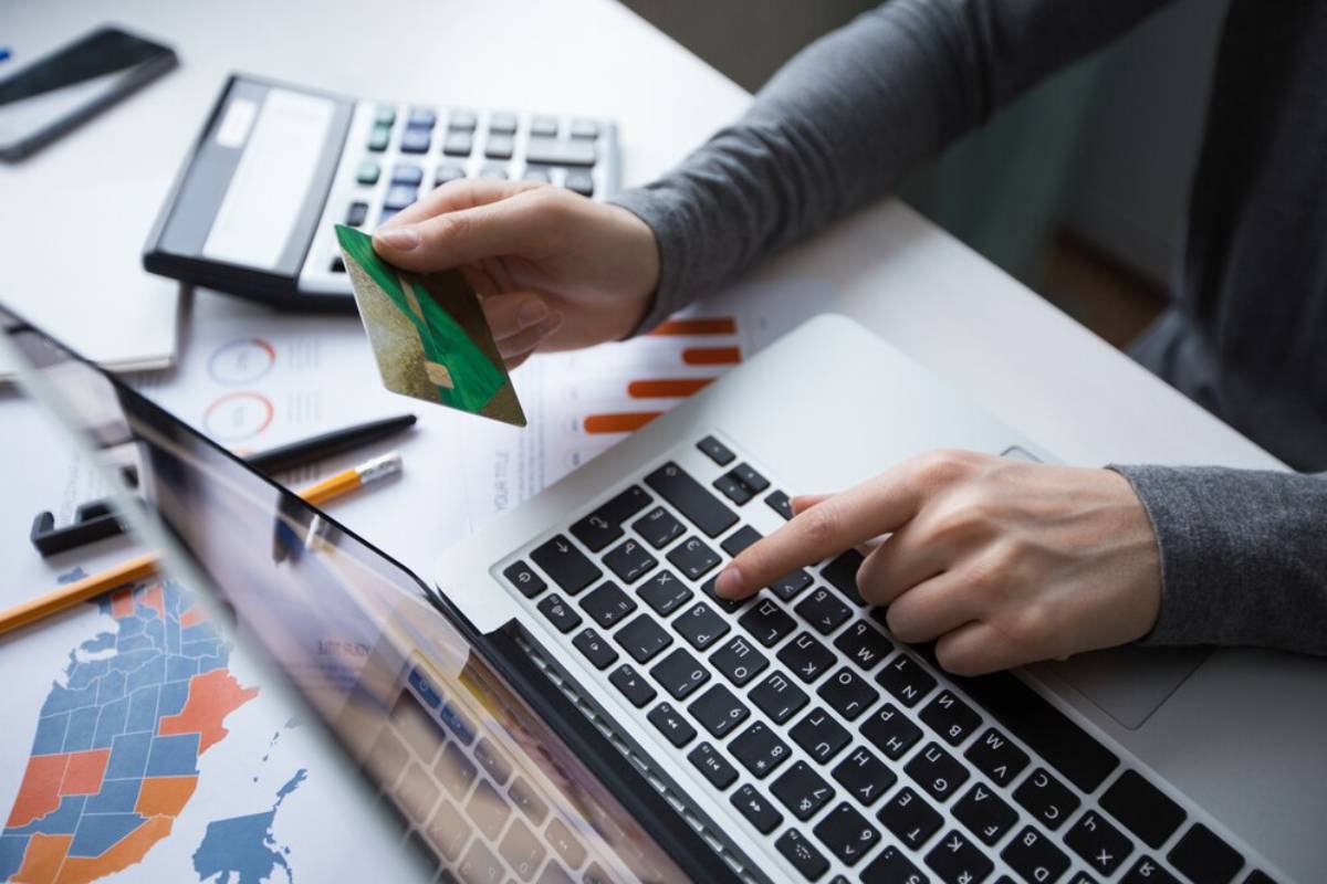 A person holding a credit card over a laptop, focusing on the keyboard while surrounded by office supplies and financial documents.