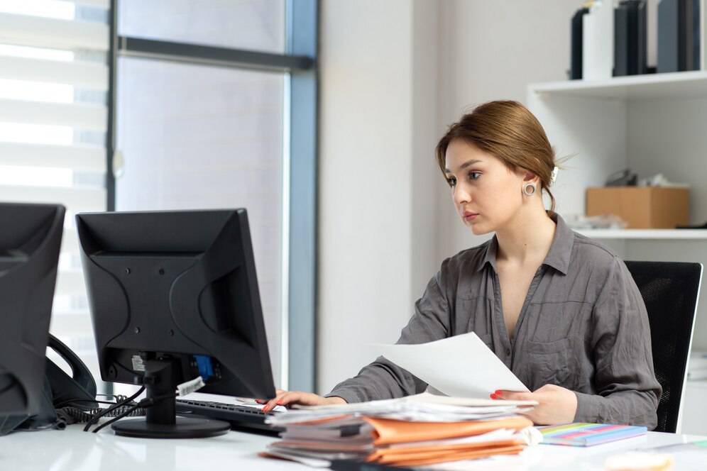 A person in a gray shirt sits at a desk, reviewing documents while using a computer in a bright, modern office environment.