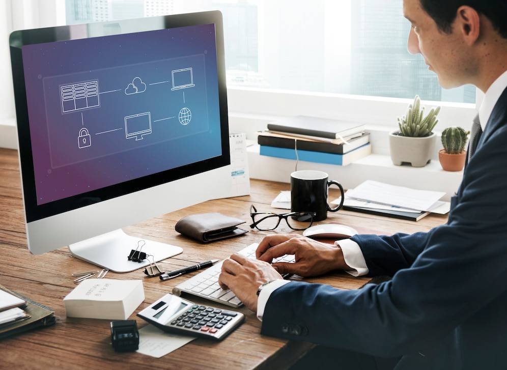 A person in a suit types on a keyboard at a desk with a computer displaying a network diagram, surrounded by office supplies and plants.