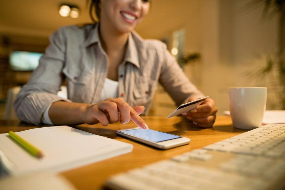 A person is using a smartphone while holding a credit card, with a cup and keyboard visible on a desk.