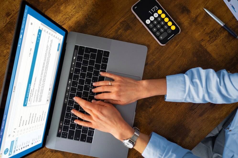 A person types on a laptop while a calculator app is visible on their phone, with a spreadsheet open on the laptop screen.