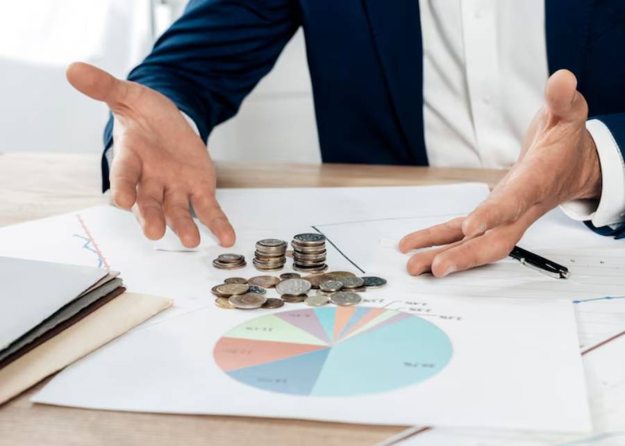 A man in a suit gestures over a table with coins and financial graphs, emphasizing a discussion about money management.