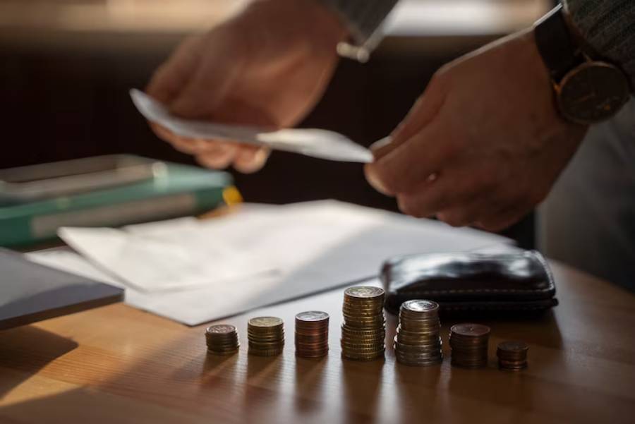 A person's hands counting cash next to a stack of coins on a wooden table with papers and a wallet in the background.