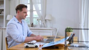 A person sits at a wooden desk using a laptop, surrounded by a potted plant, stationery, and a toy car, in a bright room.