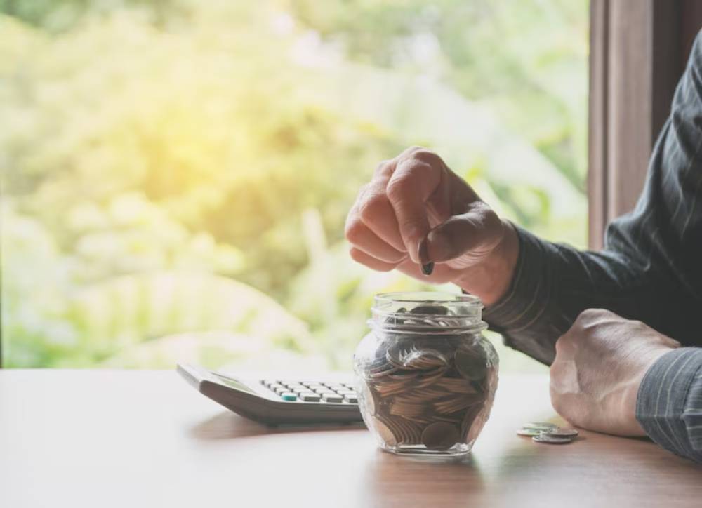 A person placing a coin into a glass jar full of coins, with a calculator nearby and greenery visible through a window.