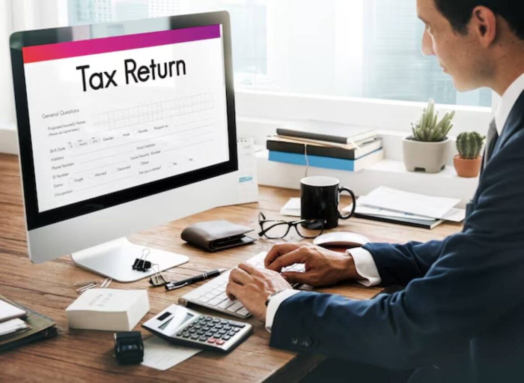 A person in a suit is working at a desk with a computer, completing a tax return form on the screen amid office supplies and a plant.