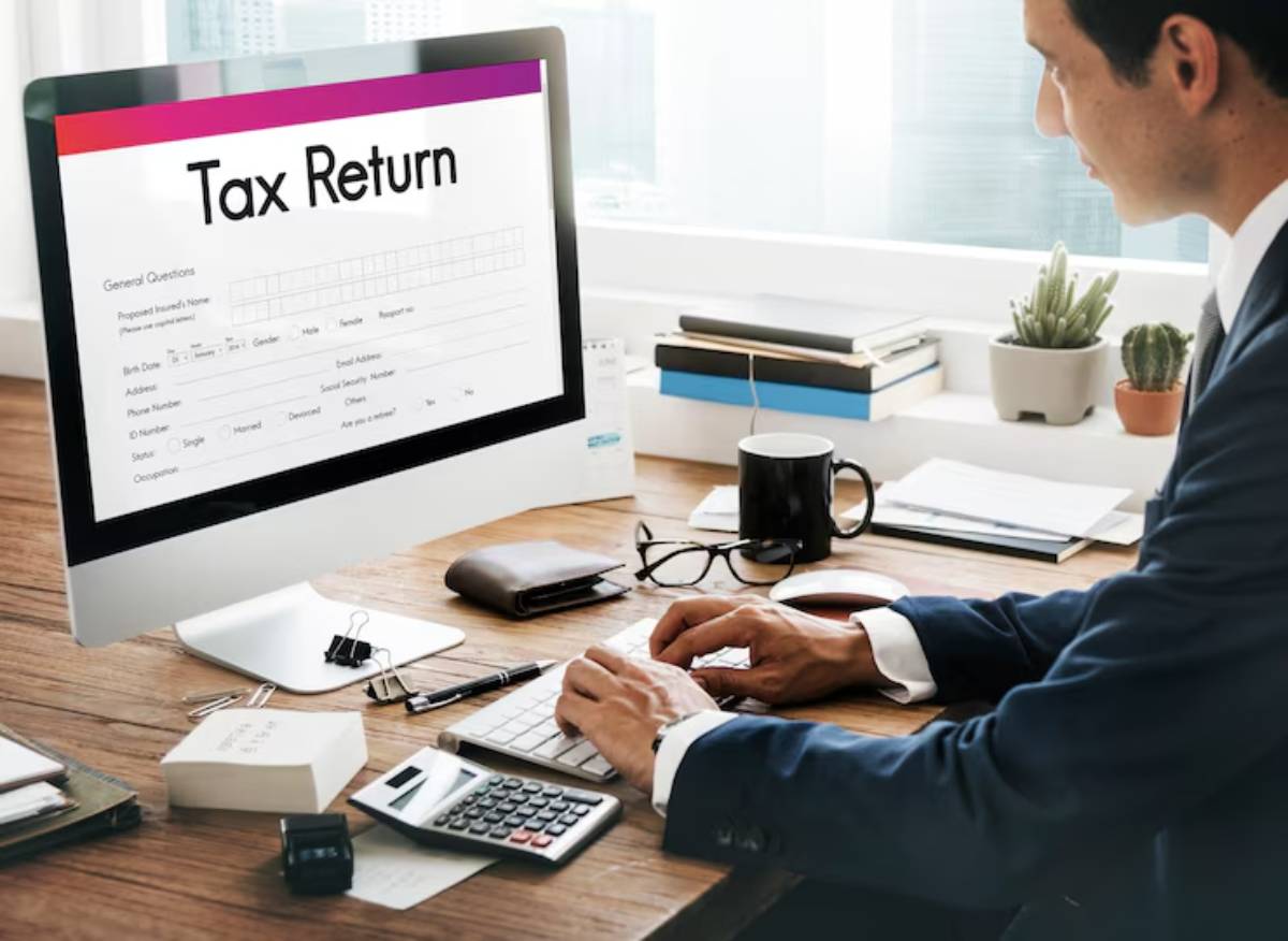 A person in a suit working at a desk, typing on a computer displaying a tax return form, with office supplies around.