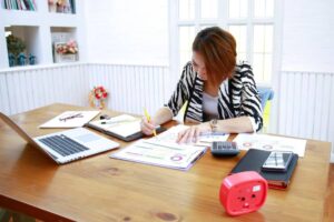 A person writes notes and analyzes documents at a wooden desk, using a laptop, calculator, and smartphone in a bright, organized space.