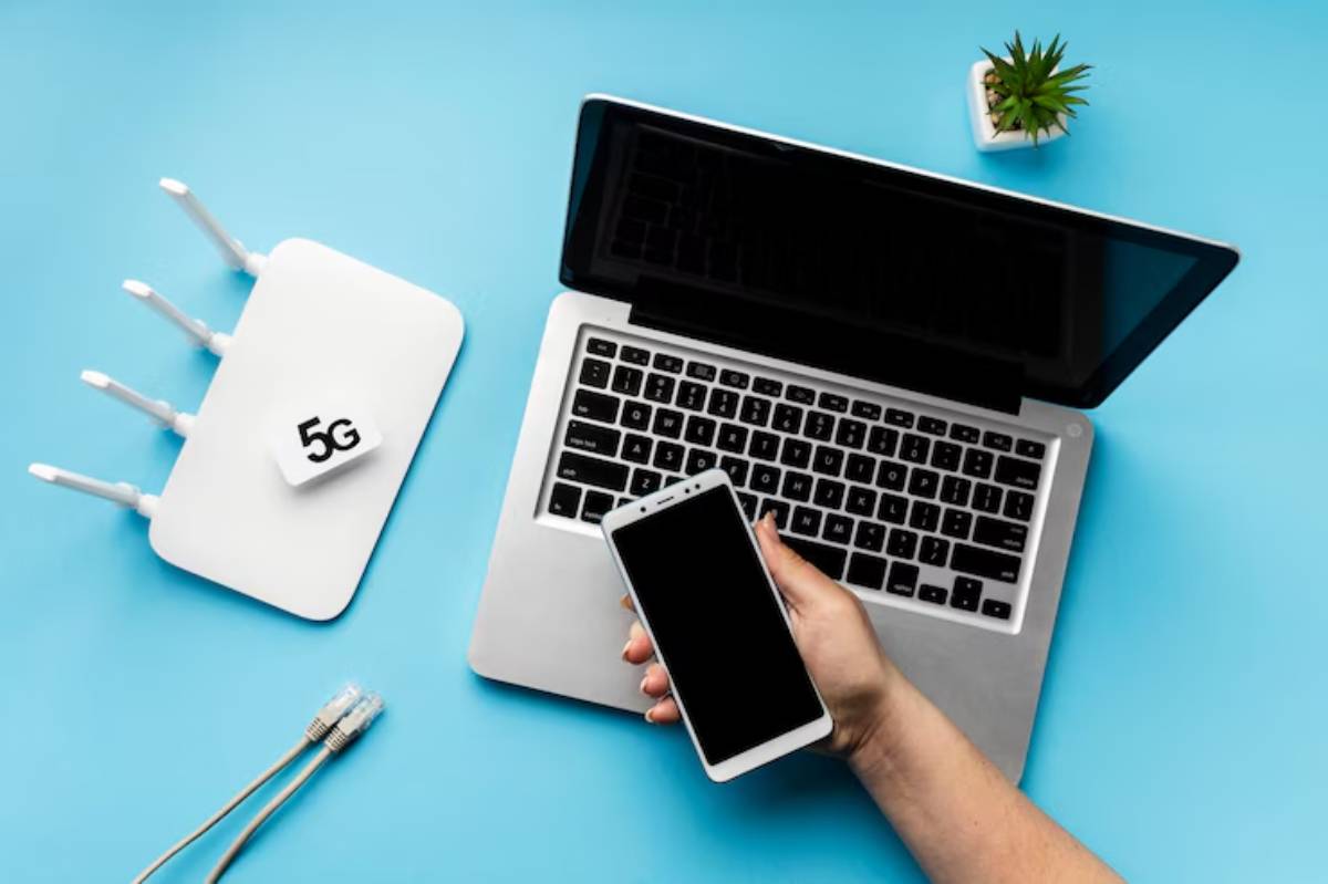  A hand holds a smartphone near a laptop and a 5G router on a blue background, with Ethernet cables beside them.