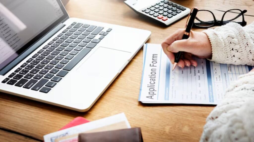 A person fills out an application form on a wooden desk, with a laptop and calculator in the background.